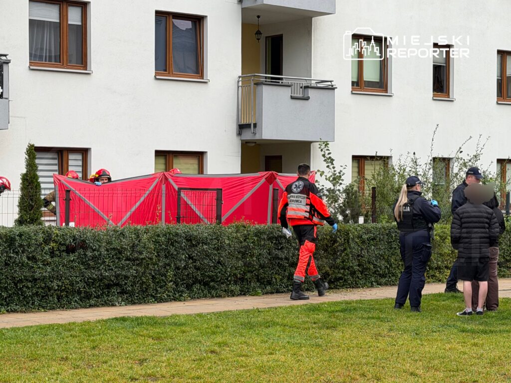 Ratownicy w odblaskowych strojach i policjanci zabezpieczają miejsce za czerwoną osłoną przy bloku mieszkalnym.