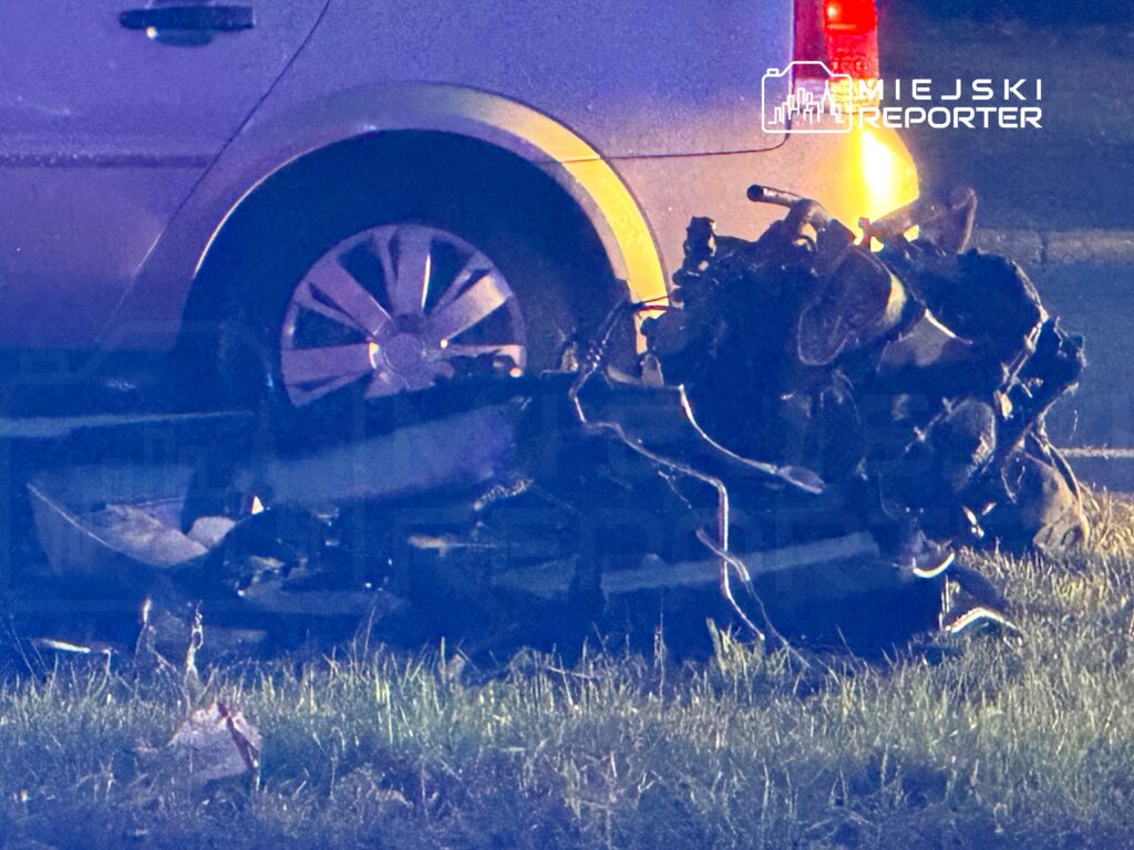 Uszkodzony motocykl leży na poboczu obok samochodu, oświetlony niebieskim i żółtym światłem nocą.