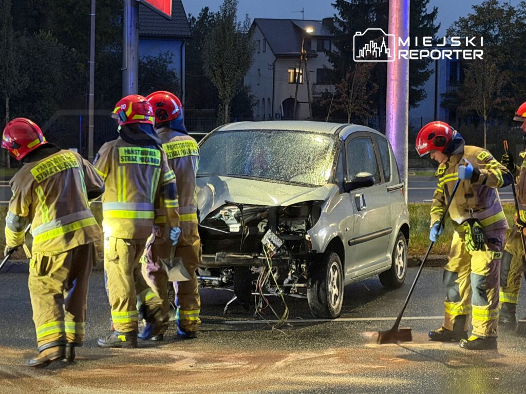 Strażacy uprzątają jezdnię obok auta z rozbitym przodem po wypadku, nocą na oświetlonej ulicy.