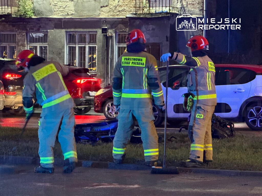 Trzech strażaków w odblaskowych uniformach zabezpiecza miejsce zdarzenia przy zaparkowanych samochodach nocą.