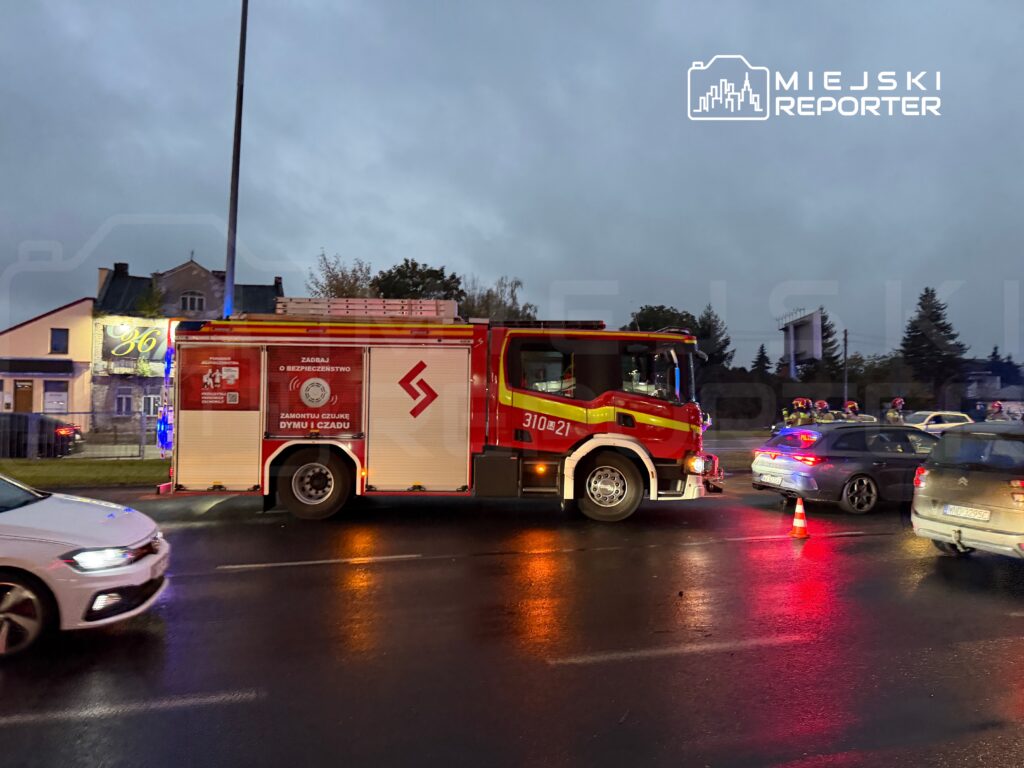 Czerwony wóz strażacki z włączonymi światłami stoi na mokrej jezdni obok samochodów i pachołka.