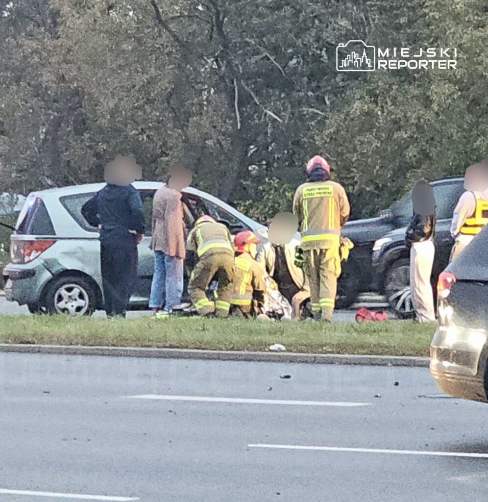 Strażacy udzielają pomocy przy dwóch samochodach po zderzeniu, na poboczu drogi otoczonym drzewami.