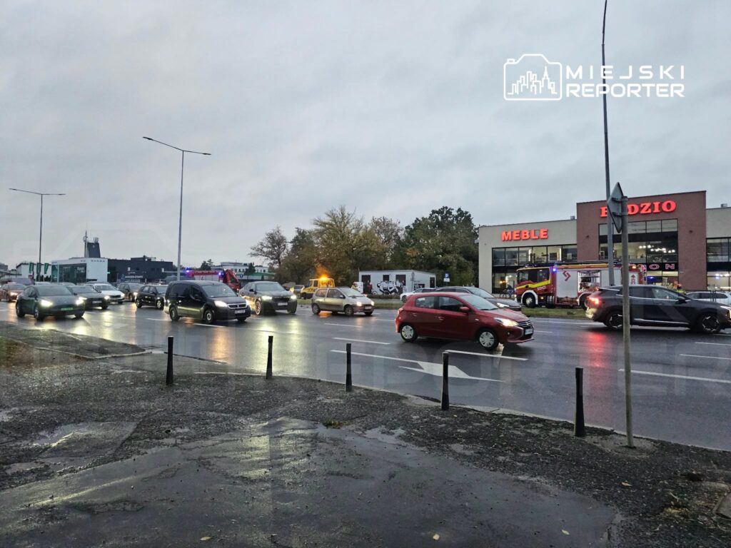 Samochody jadą po mokrej ulicy; przy sklepie meblowym stoi wóz strażacki.