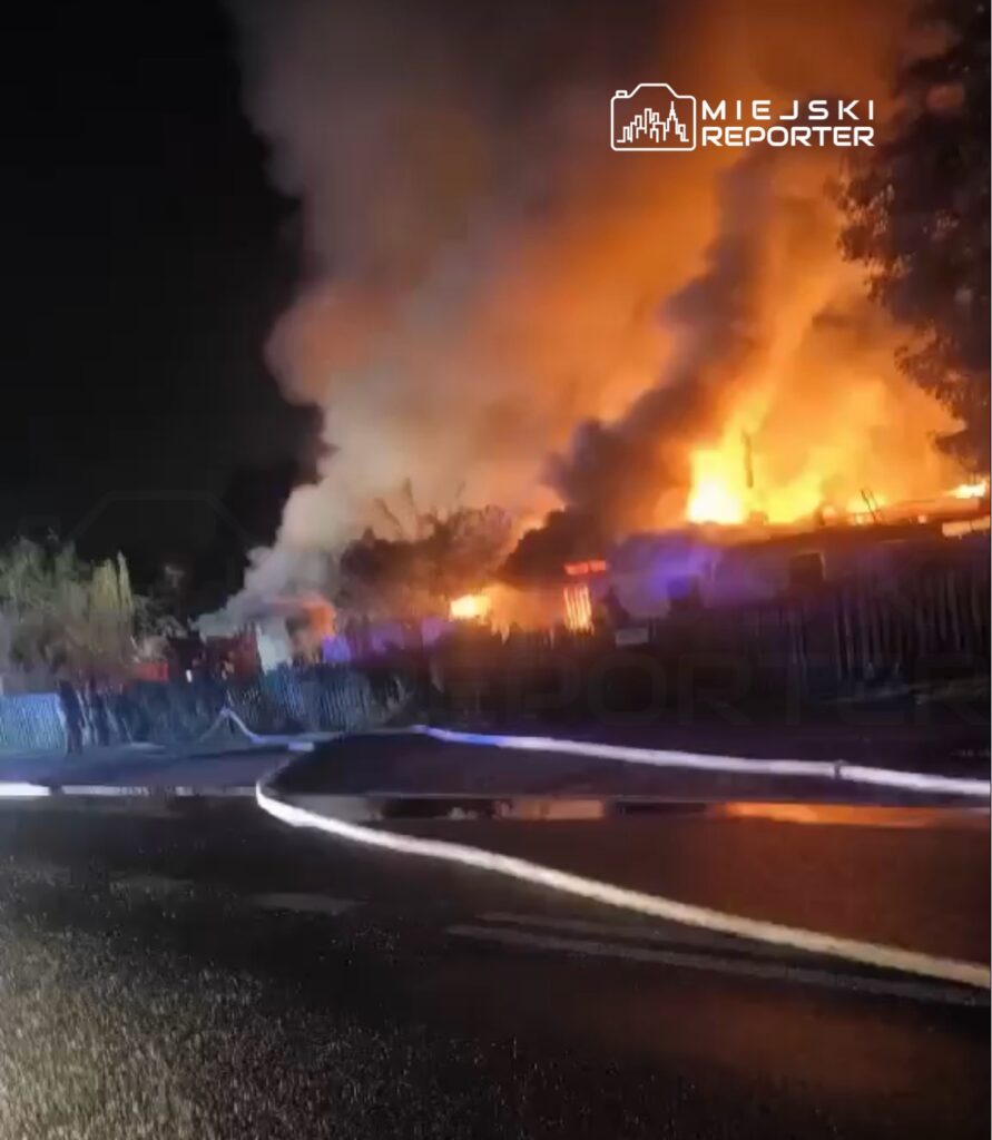 Pożar budynku nocą, płomienie i gęsty dym unoszą się za ogrodzeniem, a na jezdni leżą węże strażackie.