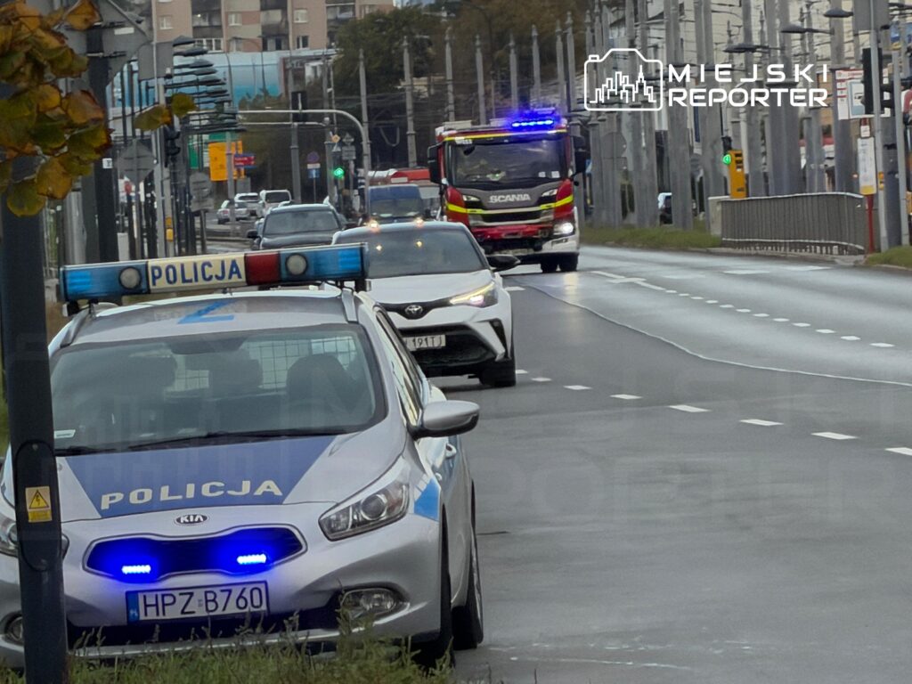 Radiowóz policji z włączonymi niebieskimi światłami stoi przy jezdni, a za nim jedzie wóz strażacki.