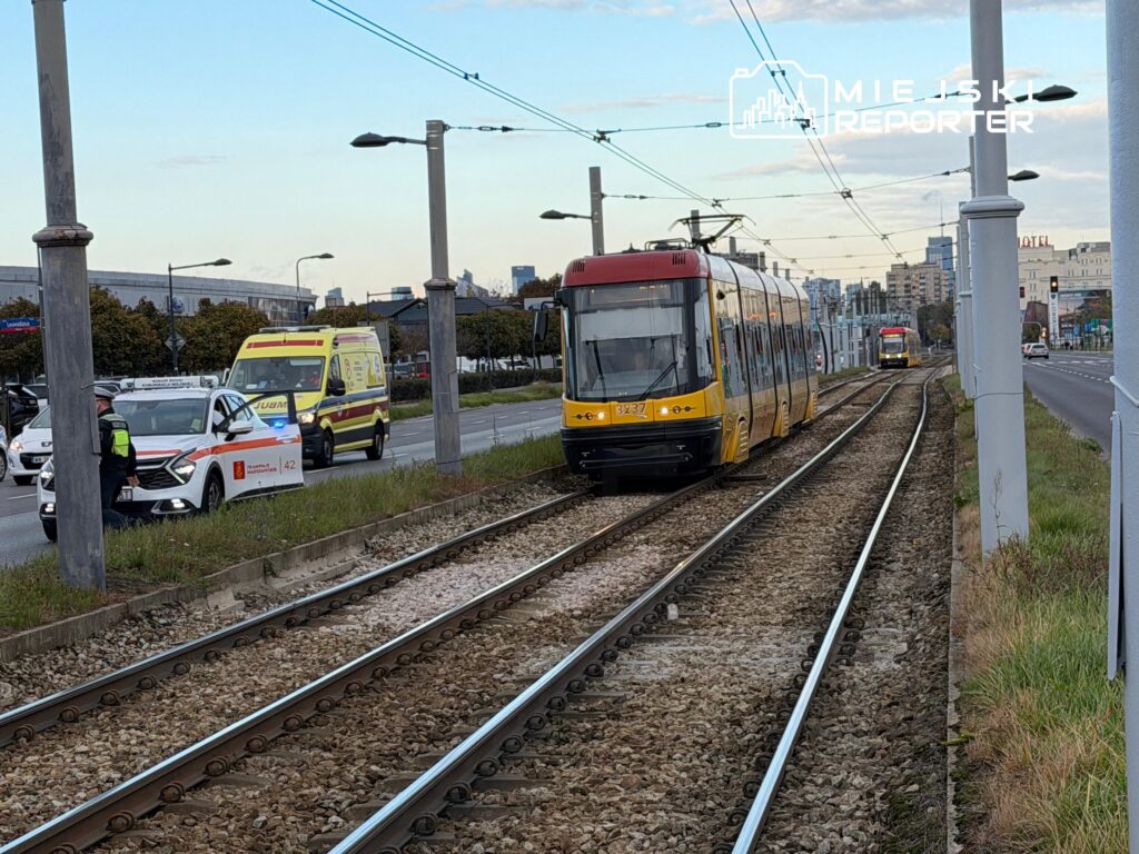 Żółto-czerwony tramwaj jedzie po torach przy ulicy, obok stoją karetka i samochód służb.