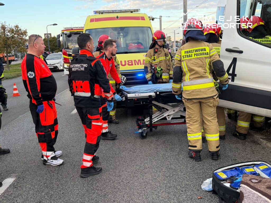 Ratownicy medyczni i strażacy przy noszach między karetką a wozem strażackim na drodze.