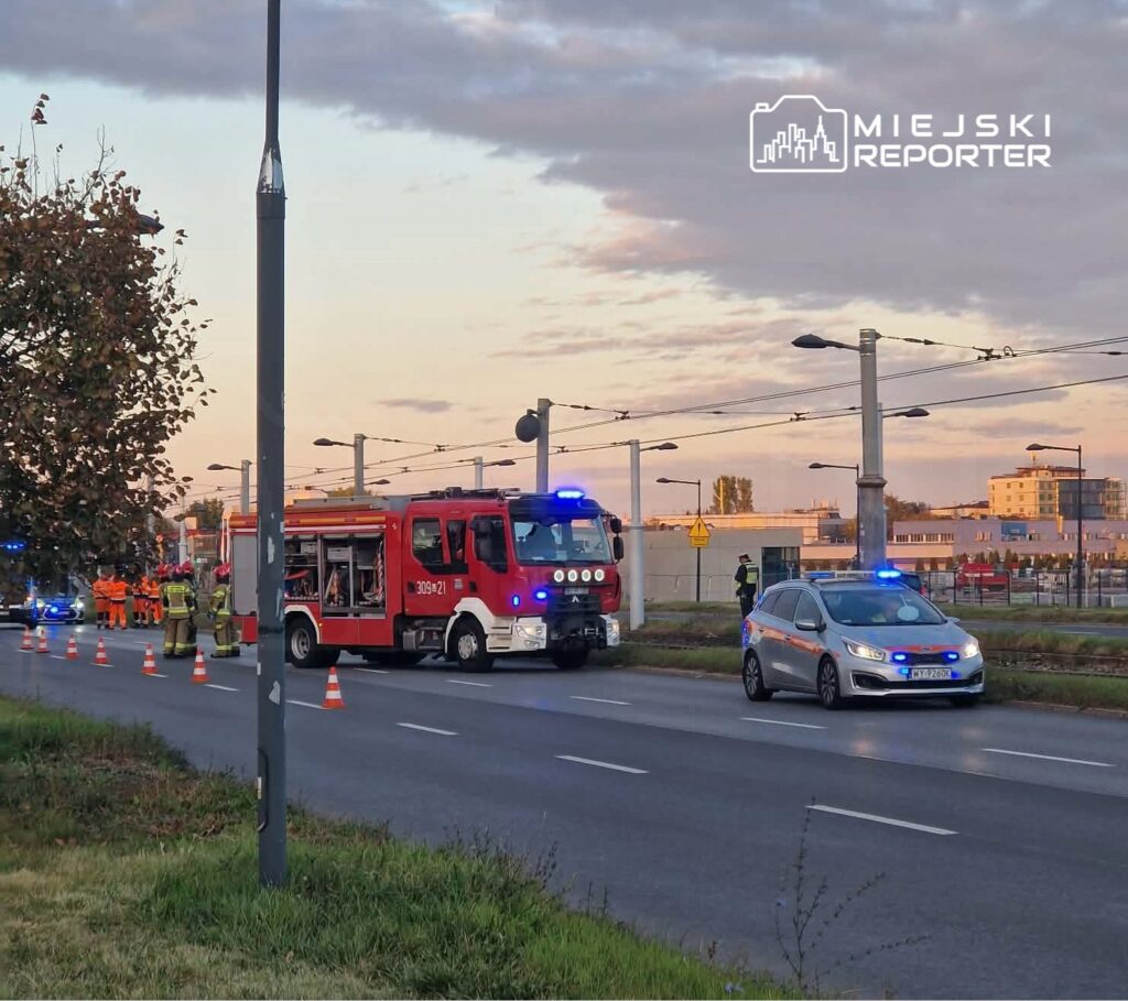 Wóz strażacki i samochód służb z włączonymi sygnałami świetlnymi stoją na drodze zabezpieczonej pachołkami.