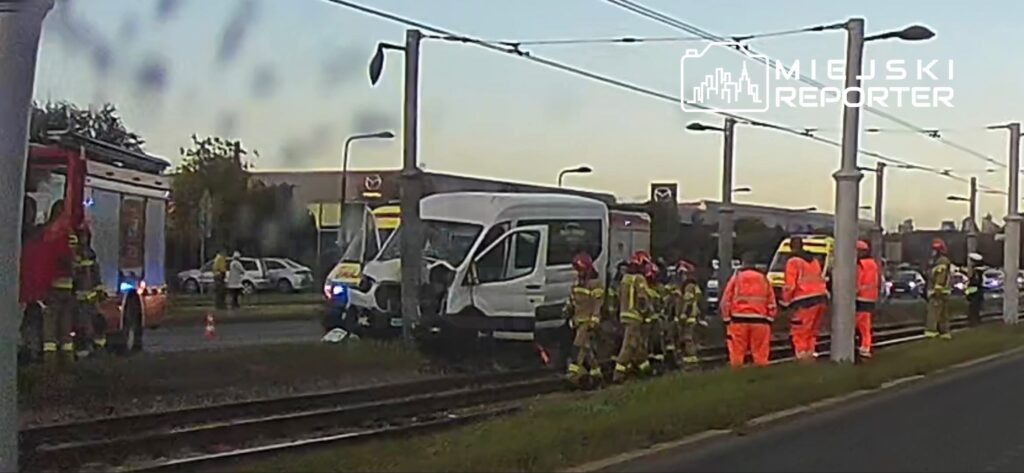 Strażacy i ratownicy przy białym busie na torowisku tramwajowym, obok stoją pojazdy służb ratunkowych.