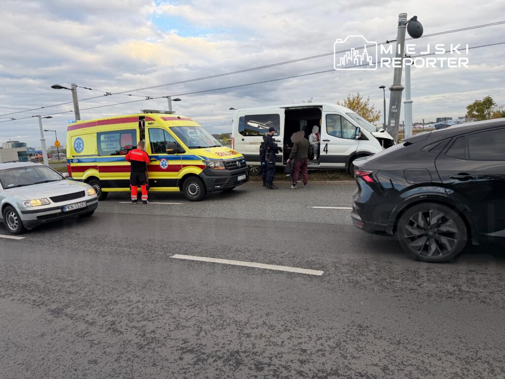 Żółty ambulans i biały bus stoją na jezdni, obok ratownik i kilka osób.