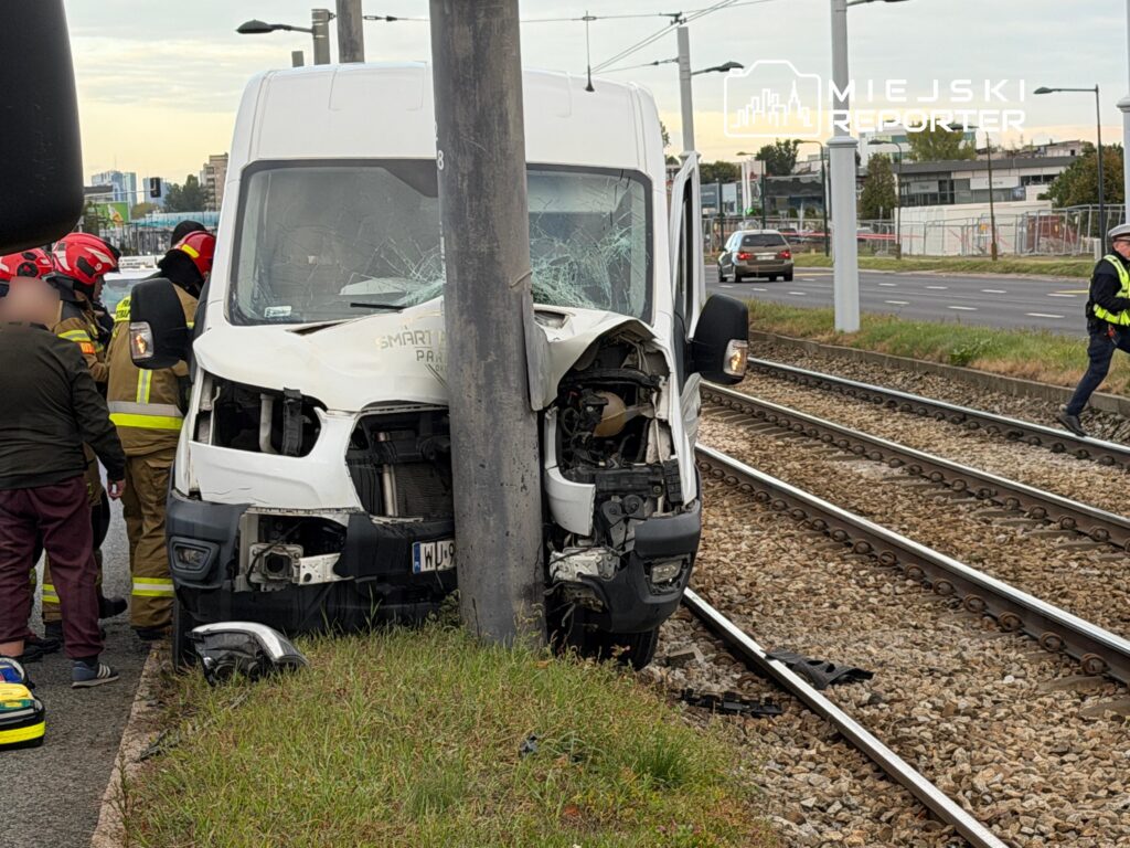 Uszkodzony biały bus stoi po zderzeniu ze słupem przy torach tramwajowych; obok pracują służby ratunkowe.