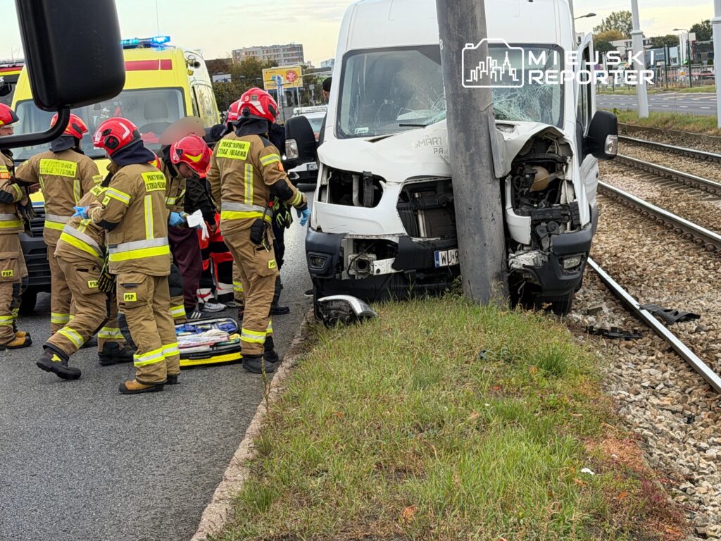 Strażacy udzielają pomocy przy rozbitym białym busie stojącym przy torach tramwajowych.