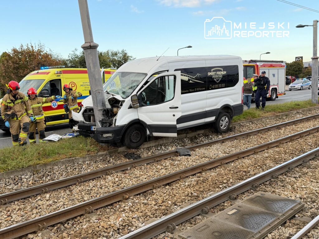 Uszkodzony biały van uderzył w słup przy torowisku; obok stoją strażacy, karetka i wóz strażacki.