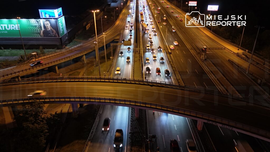 Samochody jadą nocą wielopasmową autostradą pod wiaduktem, a na sąsiednich jezdniach tworzy się korek.