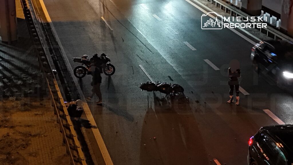 Dwa motocykle leżą na nocnej jezdni, a obok stoją osoby i samochody.