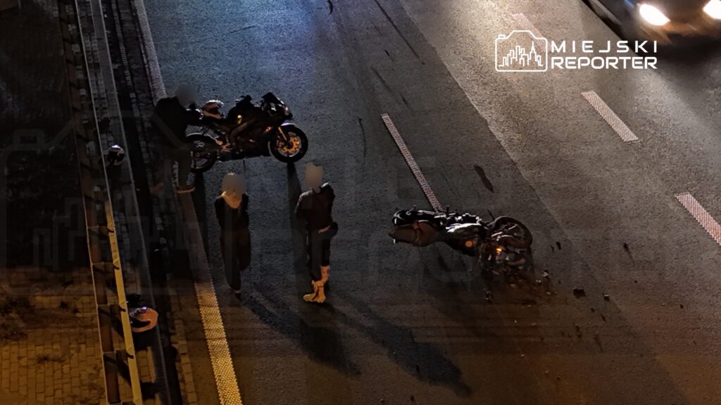 Dwa motocykle leżą na jezdni, a obok stoją dwie osoby; nocny widok z góry na ulicę.
