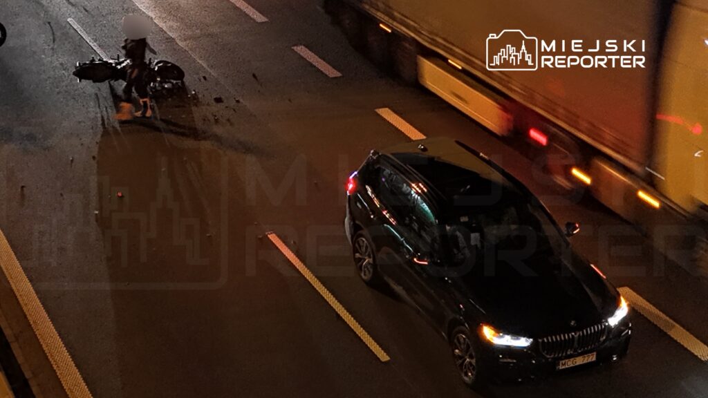 Czarny samochód osobowy i ciężarówka na nocnej jezdni, obok leżącego motocykla.