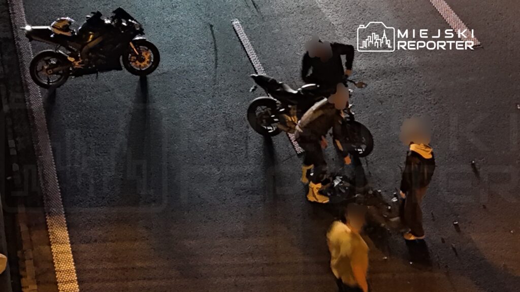 Kilka osób stoi przy dwóch motocyklach na jezdni, obok jednego z nich leży osoba.