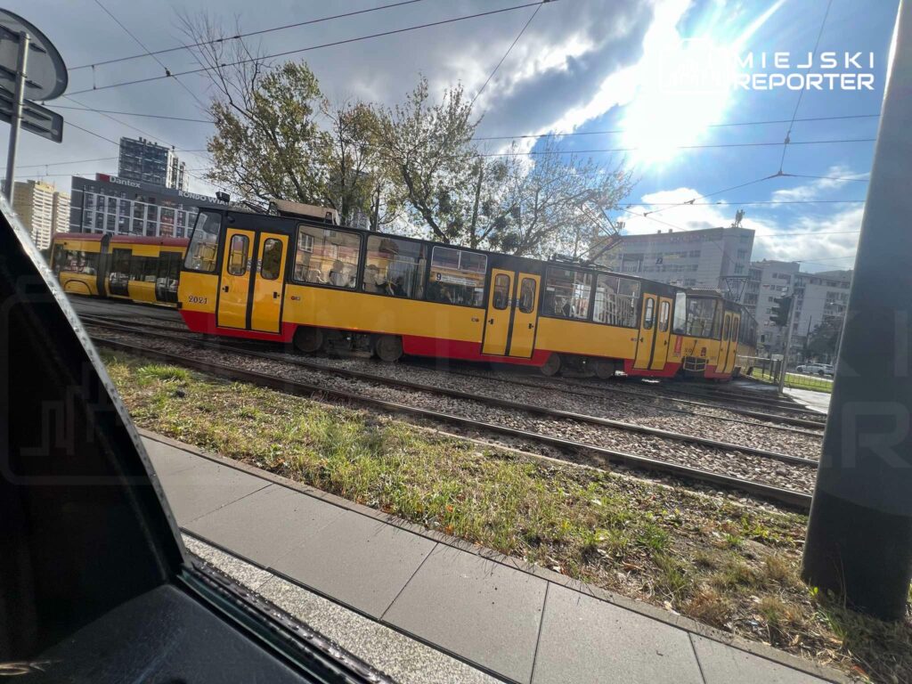 Żółto-czerwony tramwaj jedzie po torach wzdłuż ulicy, między zabudowaniami i pod siecią trakcyjną.