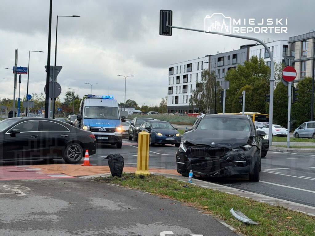 Uszkodzony czarny samochód stoi przy skrzyżowaniu, a za nim znajduje się policyjny radiowóz z włączonymi światłami.