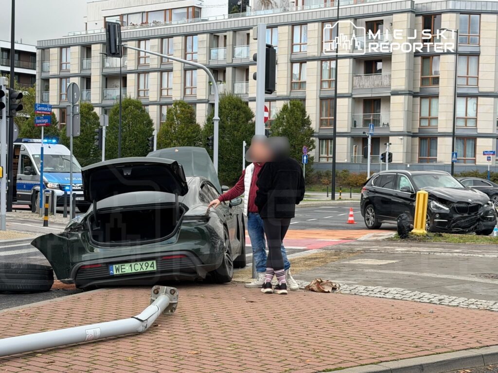 Uszkodzony samochód z otwartym bagażnikiem i osoby na miejscu kolizji na skrzyżowaniu; obok stoi radiowóz policyjny.