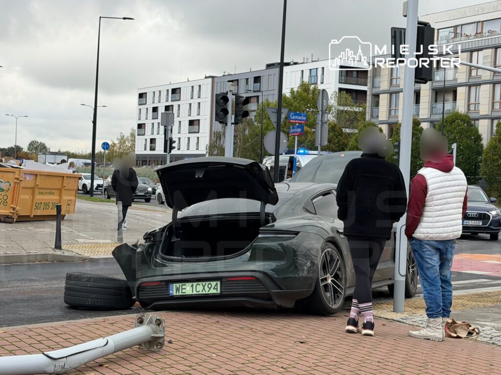 Uszkodzony sportowy samochód z otwartym bagażnikiem stoi przy skrzyżowaniu; obok leży odłączone koło i stoi kilka osób.