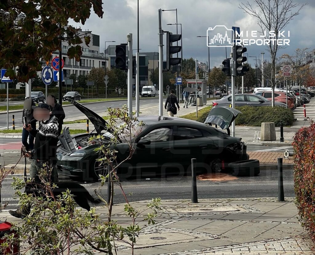 Czarny samochód z otwartą maską stoi na skrzyżowaniu, obok niego rozmawiają dwie osoby.