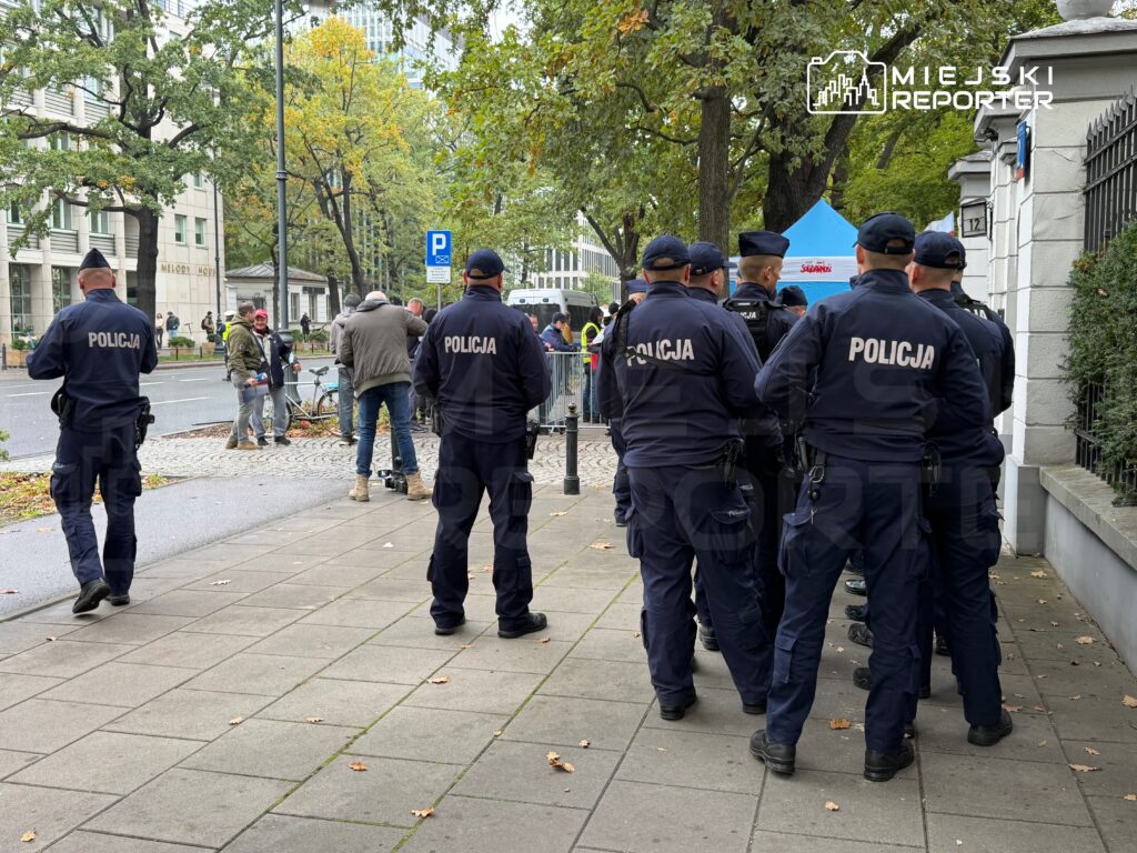 Grupa policjantów w granatowych mundurach stoi na chodniku przy ulicy, wśród drzew i miejskich budynków.