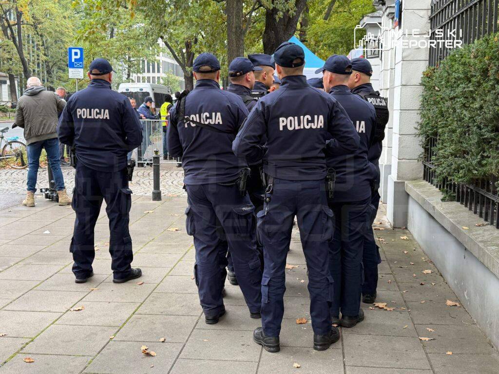 Grupa policjantów w mundurach stoi na chodniku przy ogrodzeniu i budynku, wśród drzew.