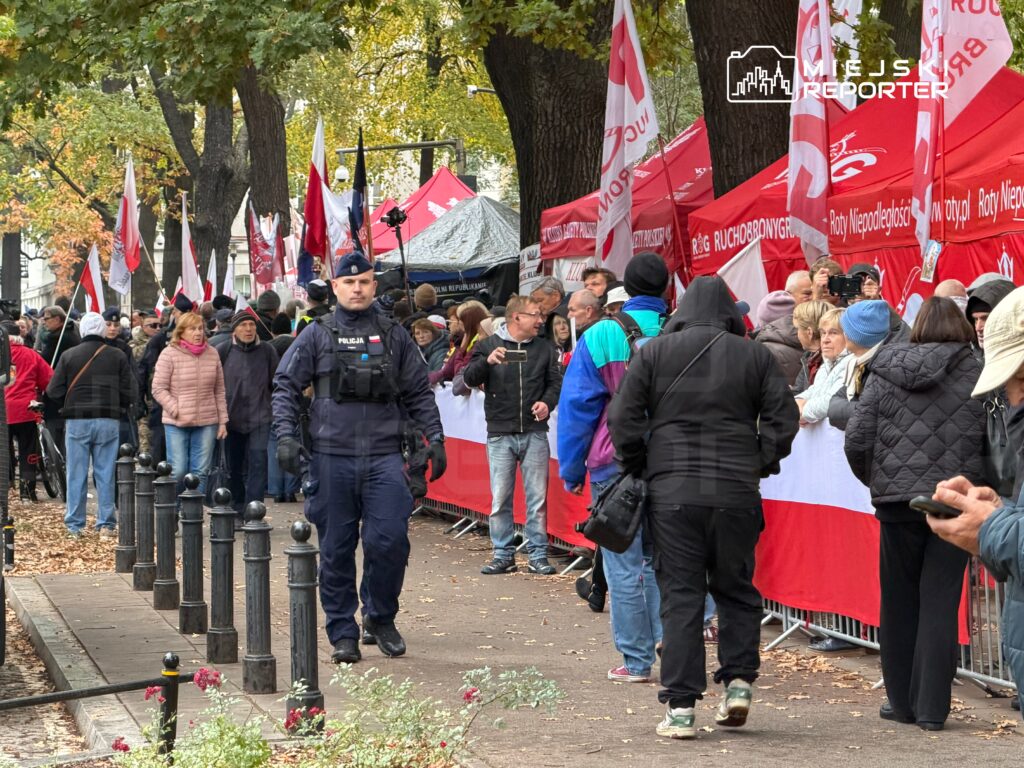 Policjant patroluje tłum ludzi przy czerwonych namiotach i flagach na zadrzewionej ulicy.