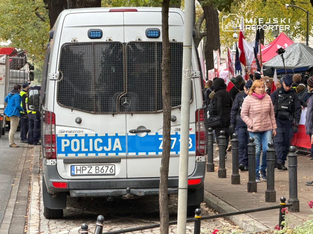 Tył policyjnego furgonu z napisem „POLICJA” na ulicy, obok zgromadzonego tłumu i namiotów z flagami.