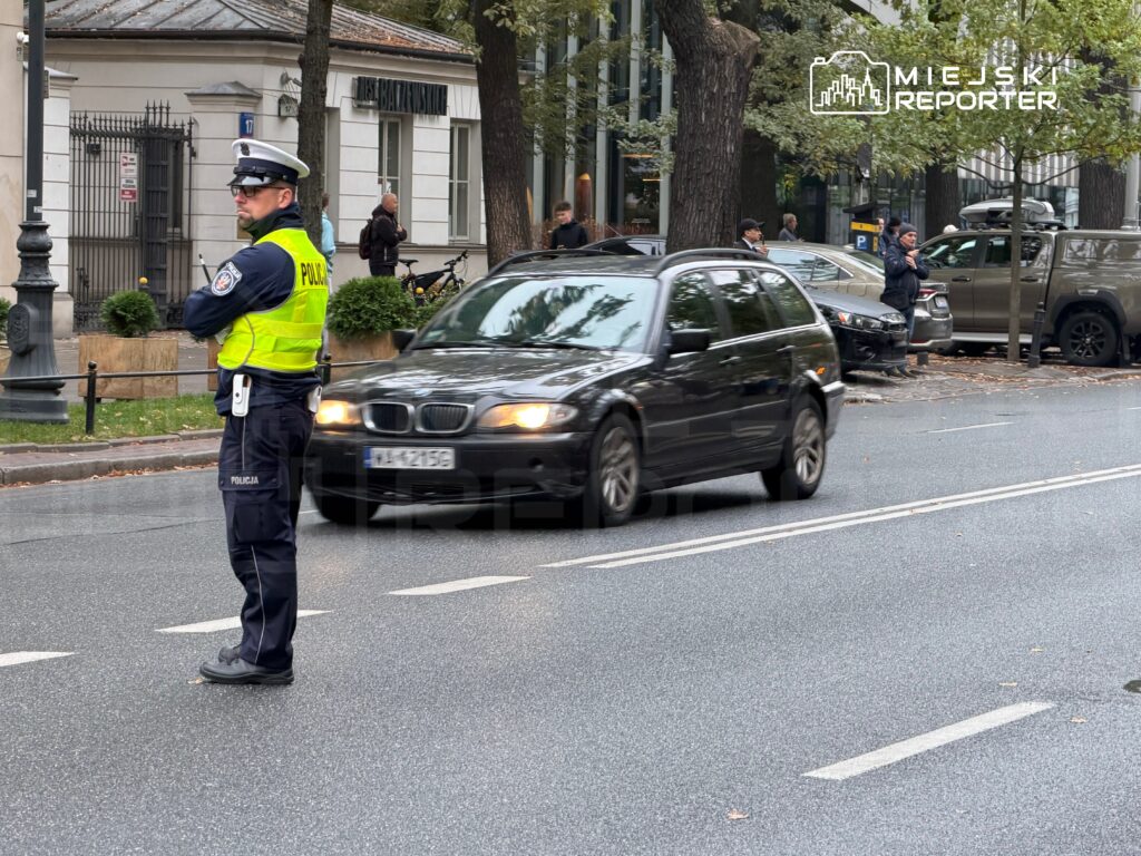 Policjant w odblaskowej kamizelce kieruje ruchem na ulicy, a obok przejeżdża samochód osobowy.