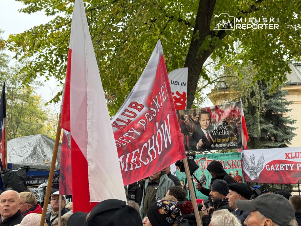 Uczestnicy zgromadzenia trzymają polskie flagi i transparenty, w tym „Klub Gazety Polskiej”, pod drzewami.