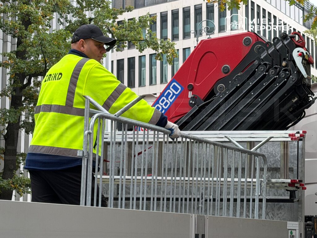 Pracownik w odblaskowej kurtce obsługuje czerwony podnośnik koszowy przy metalowym ogrodzeniu na tle biurowca.