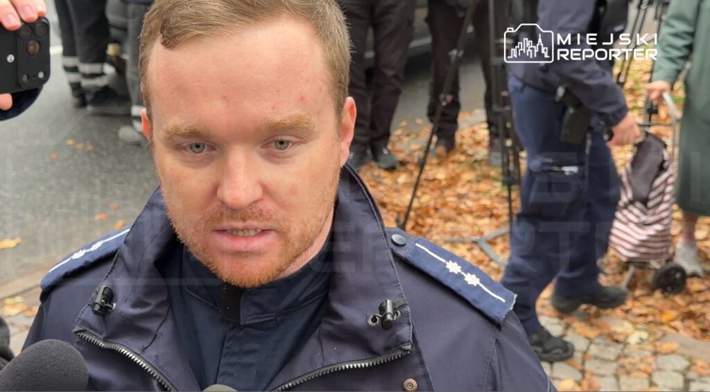 Policjant w granatowym mundurze udziela wywiadu na ulicy, w tle stoją ludzie i leżą jesienne liście.