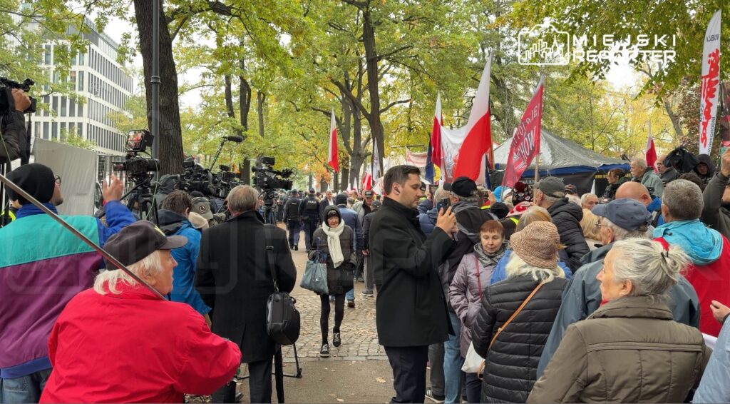 Tłum ludzi z czerwonymi i biało-czerwonymi flagami oraz kamerami gromadzi się na alei wśród drzew.