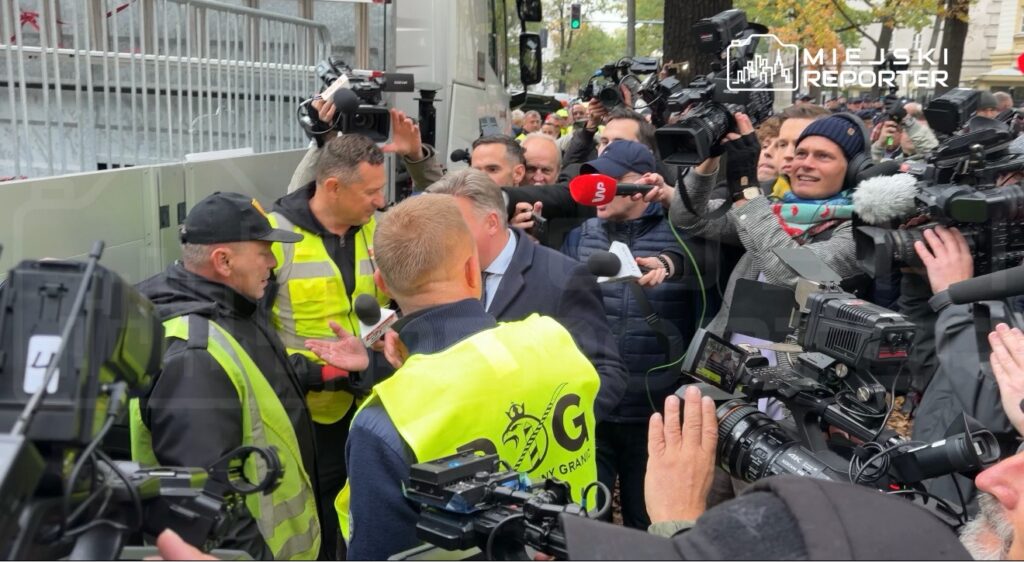 Mężczyzna w żółtej kamizelce rozmawia z policjantami, otoczony przez reporterów z kamerami i mikrofonami.