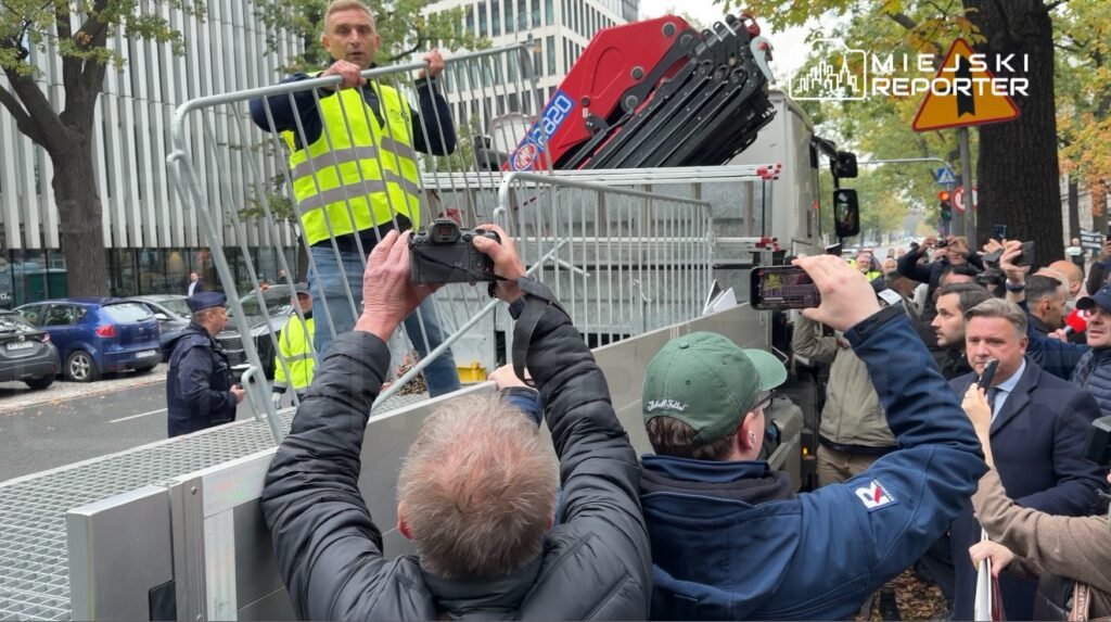 Mężczyzna w odblaskowej kamizelce montuje metalowe barierki przy ciężarówce, a tłum obserwuje i filmuje go z chodnika.
