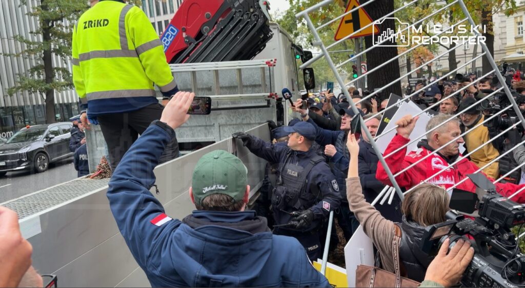 Policjanci i fotoreporterzy zgromadzeni przy ciężarówce, na której pracownik w odblaskowej kurtce obsługuje dźwig.