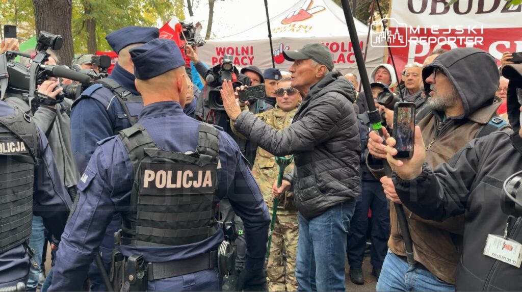 Mężczyzna w czarnej kurtce gestykuluje przed policjantami w tłumie osób z kamerami i telefonami.