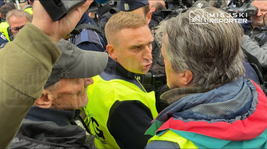 Funkcjonariusz policji w żółtej kamizelce rozmawia z mężczyzną w czerwonej kurtce w tłumie policjantów i reporterów.