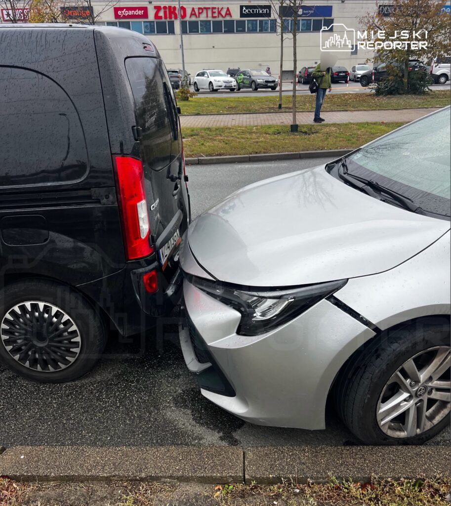 Srebrny samochód osobowy uderzył przodem w tył czarnego vana na mokrym parkingu.