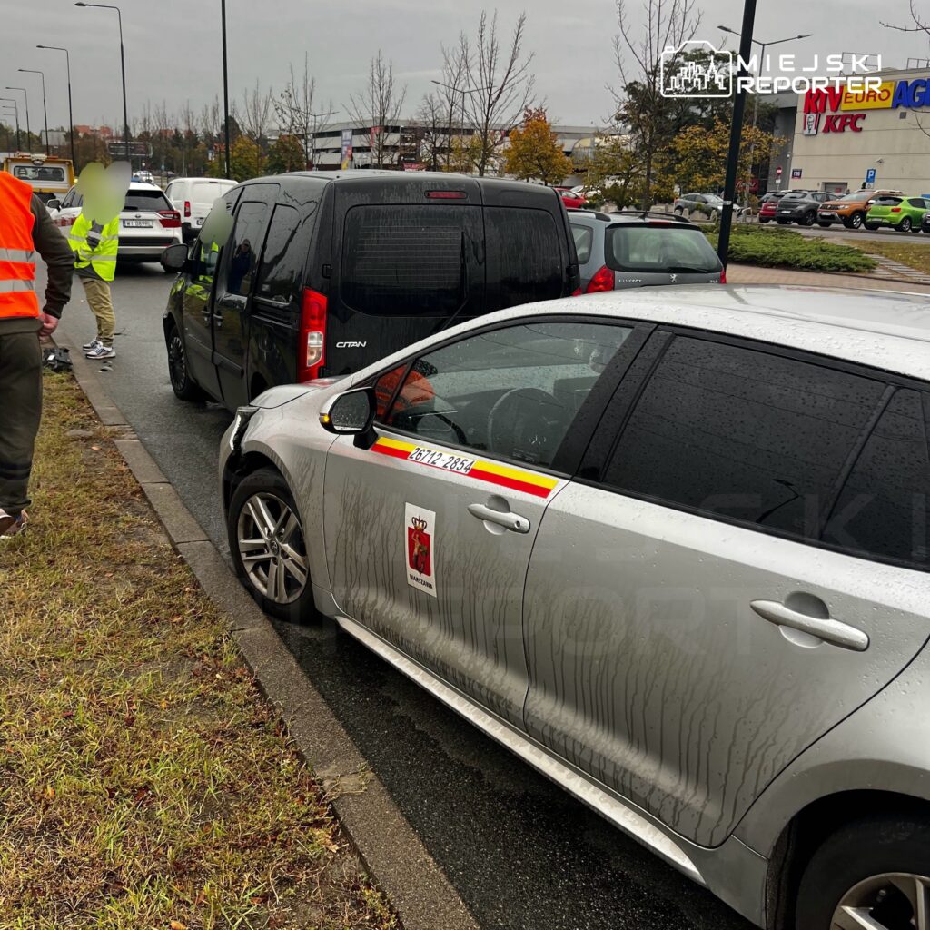 Srebrny samochód służbowy i czarny van stoją na parkingu, obok pracują osoby w kamizelkach odblaskowych.