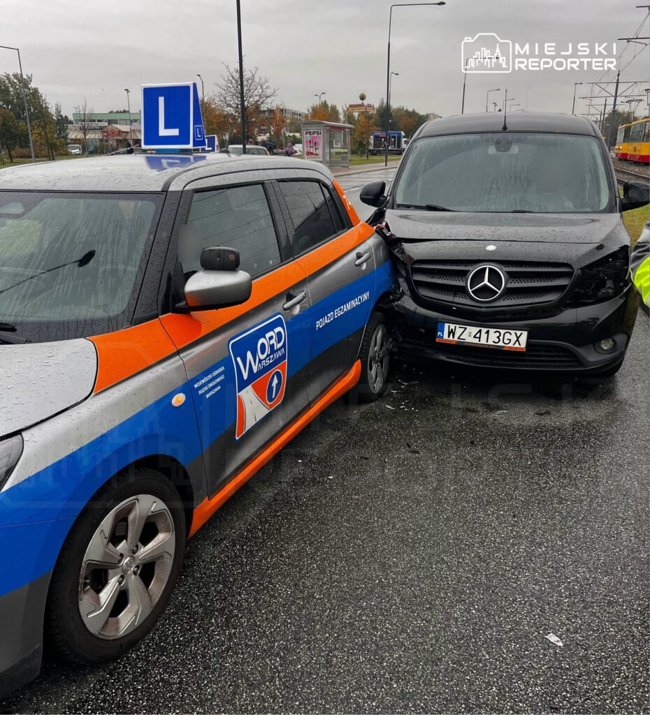 Czarny bus uderzył w samochód nauki jazdy na mokrej ulicy; pojazdy stoją obok torowiska tramwajowego.