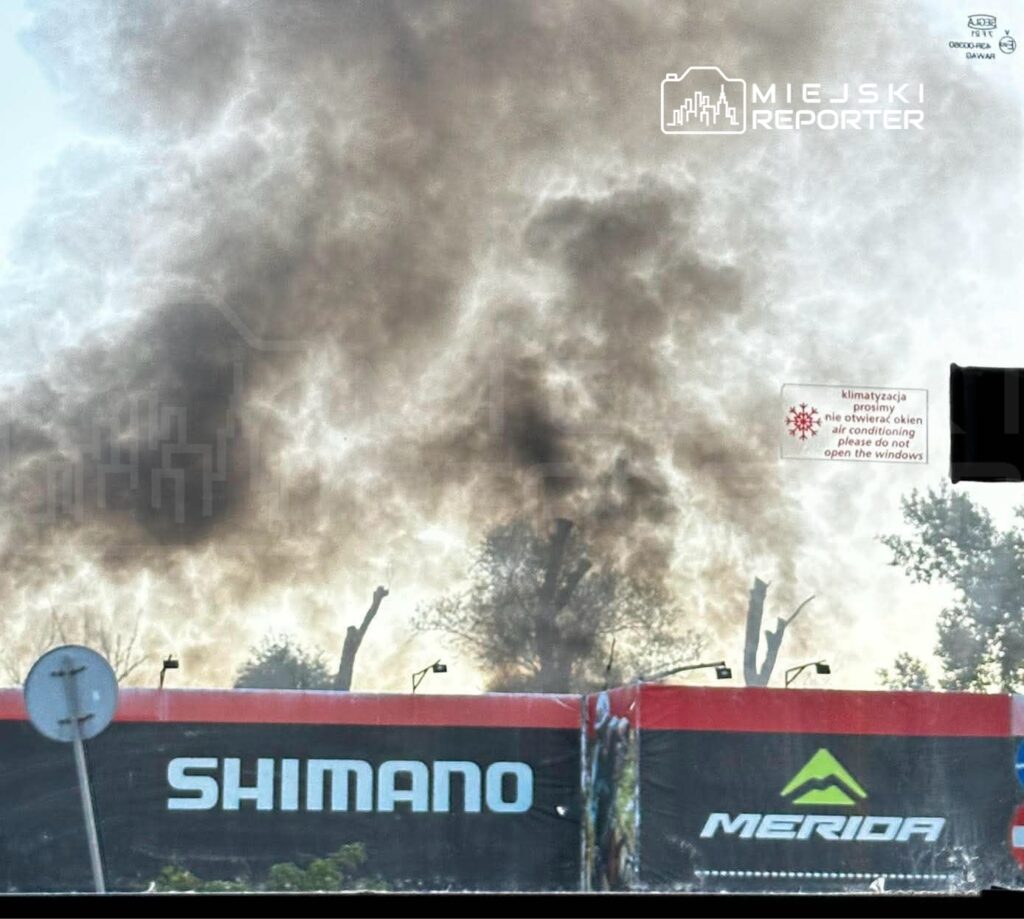 Gęsty dym unosi się za ogrodzeniem z reklamami Shimano i Merida, przy ulicy otoczonej drzewami.