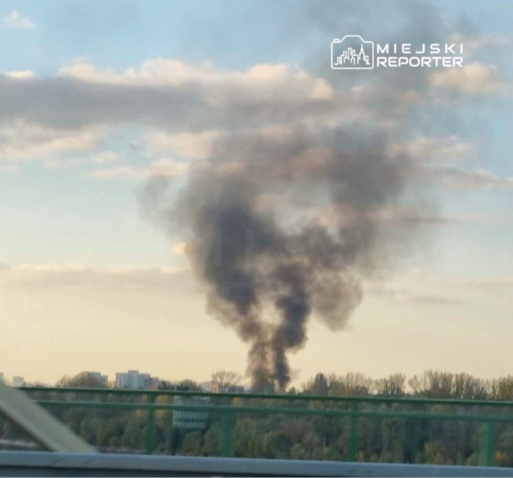 Gęsty, ciemny słup dymu unosi się nad zadrzewionym terenem i zabudowaniami, widok zza bariery mostu.