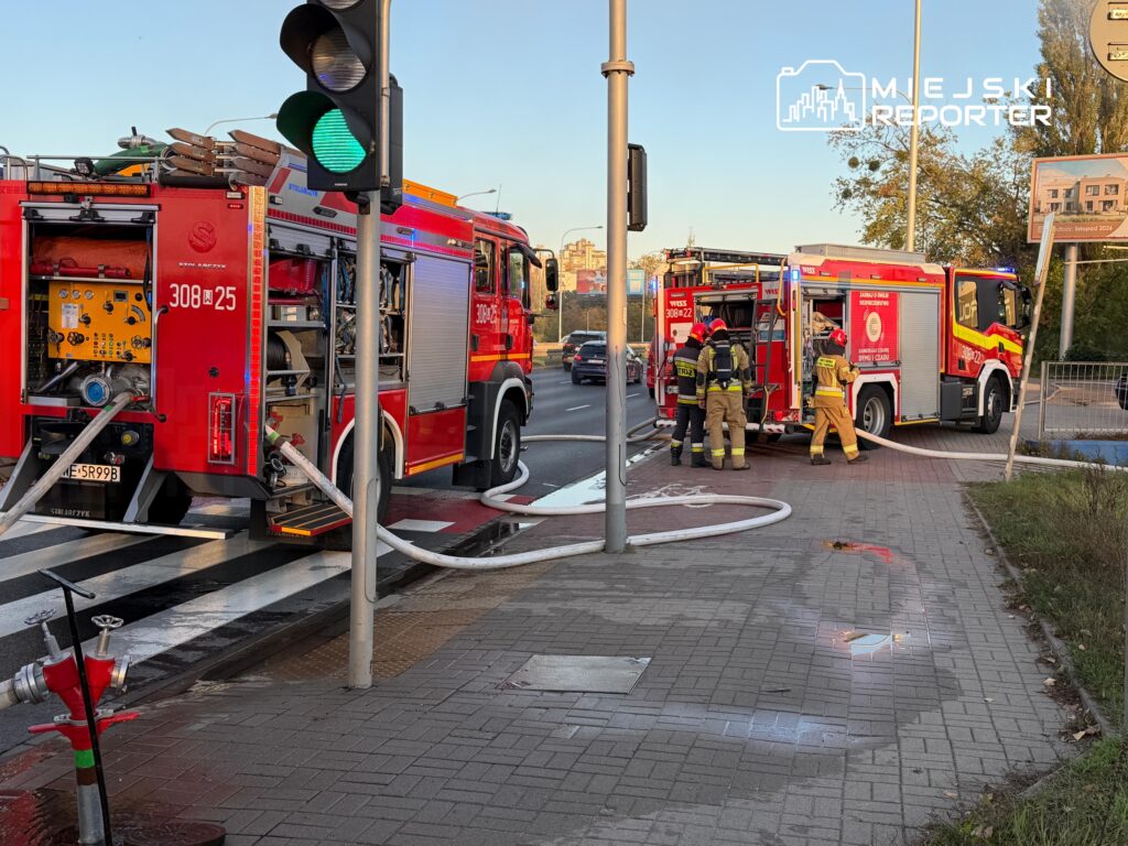 Strażacy przy dwóch czerwonych wozach pożarniczych rozstawiają sprzęt i węże na skrzyżowaniu ulic.