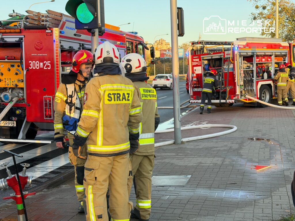 Strażacy w ubraniach ochronnych stoją przy wozach strażackich na skrzyżowaniu, obok rozłożonego węża gaśniczego.