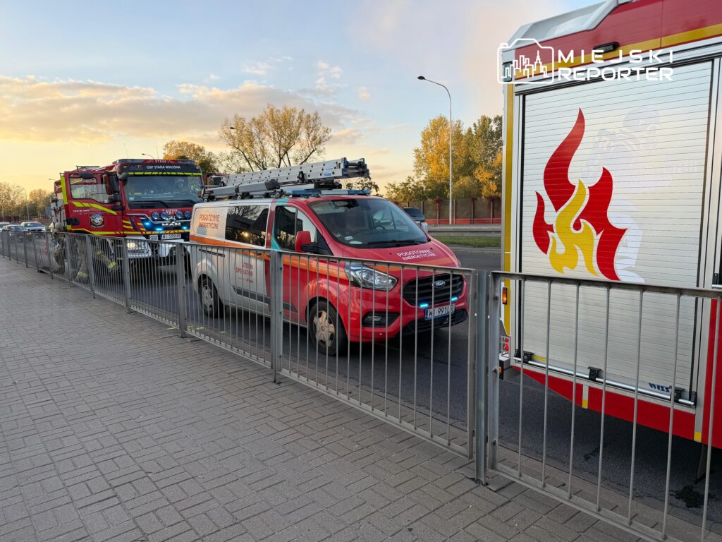 Czerwone wozy strażackie i samochód ratowniczy ustawione za metalowym ogrodzeniem przy ulicy o zmierzchu.