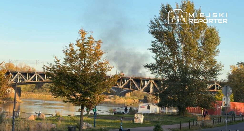 Dym unosi się nad stalowym mostem nad rzeką, widocznym za drzewami w jesiennym otoczeniu.